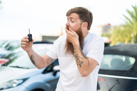 Téléchargez les photos : Homme rousse avec barbe tenant des clés de voiture à l'extérieur avec une expression faciale surprise et choquée - en image libre de droit