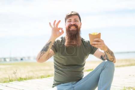 Foto de Pelirrojo con barba sosteniendo papas fritas al aire libre mostrando signo de ok con los dedos - Imagen libre de derechos