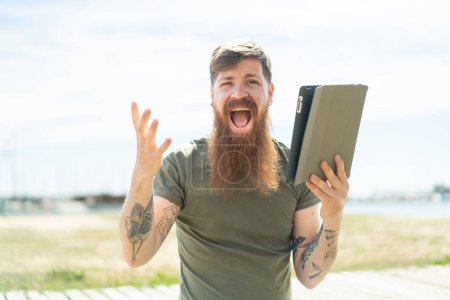 Téléchargez les photos : Homme rousse avec barbe tenant une tablette à l'extérieur avec une expression faciale choquée - en image libre de droit