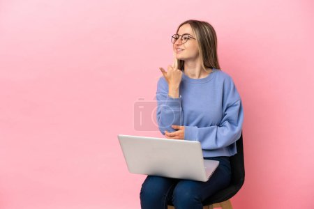 Téléchargez les photos : Jeune femme assise sur une chaise avec ordinateur portable sur fond rose isolé pointant vers le côté pour présenter un produit - en image libre de droit