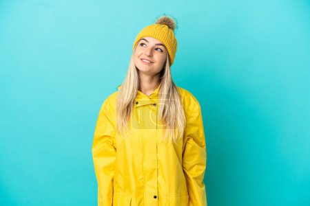 Young woman wearing a rainproof coat over isolated blue background thinking an idea while looking up