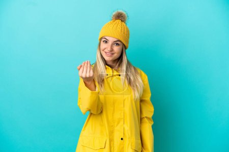 Young woman wearing a rainproof coat over isolated blue background inviting to come with hand. Happy that you came