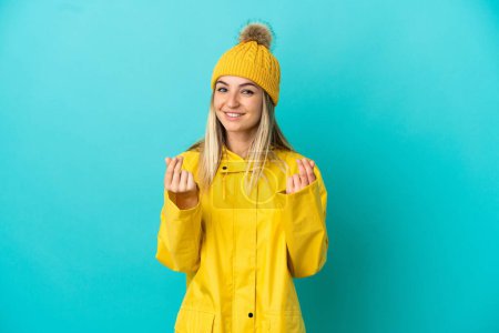 Young woman wearing a rainproof coat over isolated blue background making money gesture