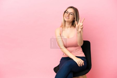 Téléchargez les photos : Jeune femme assise sur une chaise sur fond rose isolé heureux et comptant trois avec les doigts - en image libre de droit
