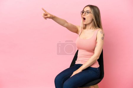 Photo for Young woman sitting on a chair over isolated pink background pointing away - Royalty Free Image