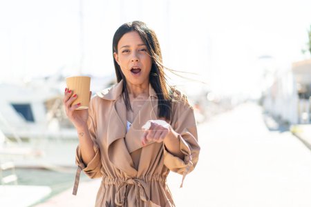 Téléchargez les photos : Jeune femme à l'extérieur tenant un café à emporter à l'extérieur surpris et pointant devant - en image libre de droit