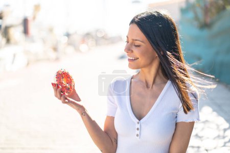 Photo for Young woman at outdoors holding a donut at outdoors with happy expression - Royalty Free Image