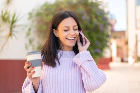 Téléchargez les photos : Jeune femme à l'extérieur en utilisant un téléphone portable et tenant un café avec une expression heureuse - en image libre de droit