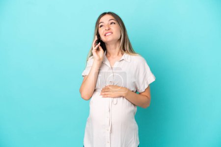 Young Romanian woman isolated on blue background pregnant and using mobile phone