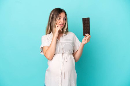Foto de Joven rumana aislada sobre fondo azul embarazada y teniendo dudas mientras sostiene chocolate - Imagen libre de derechos