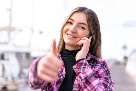 Junge hübsche Rumänin im Freien hält ein Gespräch mit dem Handy, während sie Daumen hoch macht