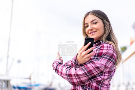 Photo for Young pretty Romanian woman at outdoors using mobile phone and pointing back - Royalty Free Image