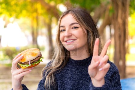 Photo for Young pretty Romanian woman holding a burger at outdoors smiling and showing victory sign - Royalty Free Image
