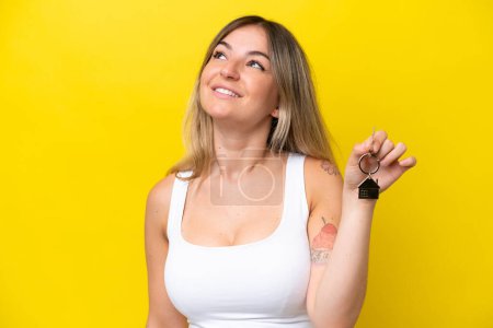 Foto de Mujer rumana joven sosteniendo llaves de casa aisladas sobre fondo amarillo mirando hacia arriba mientras sonríe - Imagen libre de derechos