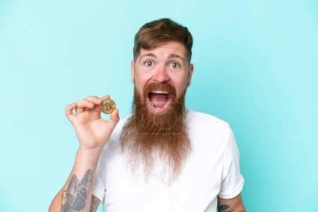 Photo for Redhead man with long beard holding a Bitcoin isolated on blue background with surprise and shocked facial expression - Royalty Free Image