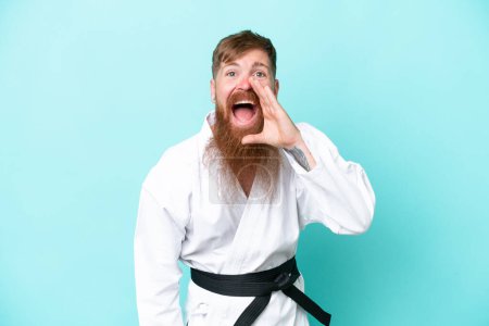 Téléchargez les photos : Homme rousse à longue barbe faisant du karaté isolé sur fond bleu criant avec la bouche grande ouverte - en image libre de droit