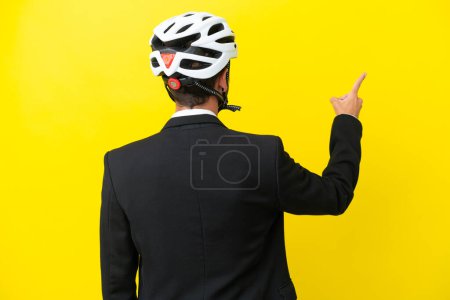 Foto de Hombre caucásico de negocios con un casco de bicicleta aislado sobre fondo amarillo apuntando hacia atrás con el dedo índice - Imagen libre de derechos
