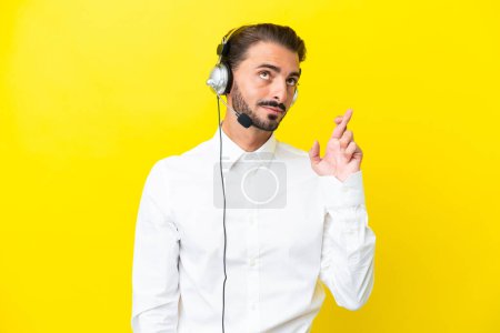 Photo for Telemarketer caucasian man working with a headset isolated on yellow background with fingers crossing and wishing the best - Royalty Free Image