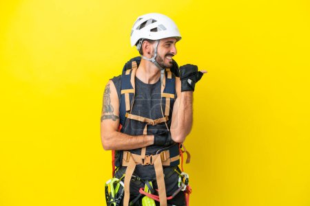 Foto de Joven escalador caucásico aislado sobre fondo amarillo apuntando hacia un lado para presentar un producto - Imagen libre de derechos