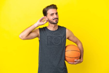 Téléchargez les photos : Beau jeune joueur de basket-ball homme isolé sur fond jaune ayant des doutes - en image libre de droit