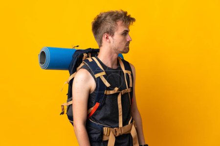 Foto de Young mountaineer man with a big backpack isolated on yellow background looking to the side - Imagen libre de derechos