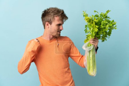 Photo for Young blonde man holding a celery isolated on blue background celebrating a victory - Royalty Free Image