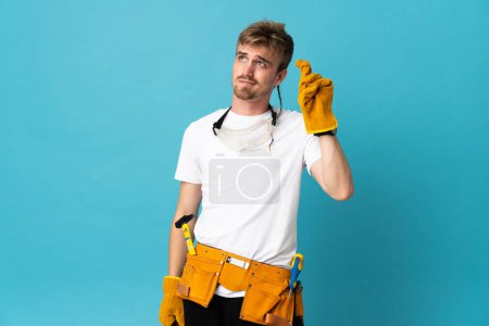 Photo for Young electrician man over isolated wall with fingers crossing and wishing the best - Royalty Free Image