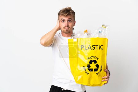 Foto de Joven hombre rubio sosteniendo una bolsa de reciclaje llena de papel para reciclar aislado sobre fondo blanco frustrado y cubriendo las orejas - Imagen libre de derechos