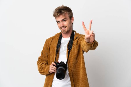 Foto de Joven fotógrafo hombre rubio aislado sobre fondo blanco sonriendo y mostrando signo de victoria - Imagen libre de derechos