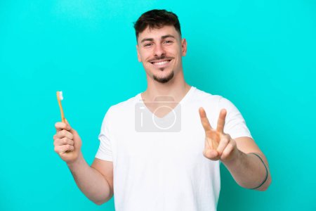 Foto de Joven hombre caucásico cepillándose los dientes aislados sobre fondo azul sonriendo y mostrando signo de victoria - Imagen libre de derechos