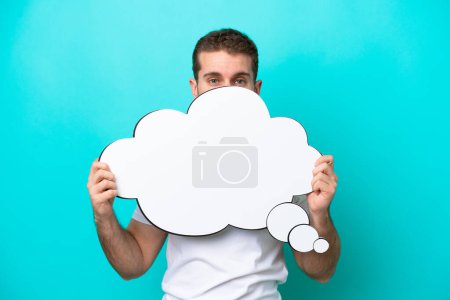 Photo for Young caucasian man isolated on blue background holding a thinking speech bubble and hiding behind it - Royalty Free Image