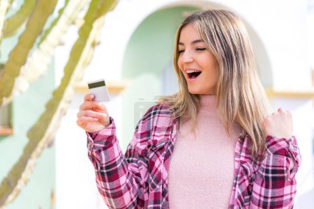 Foto de Joven mujer rumana bonita sosteniendo una tarjeta de crédito al aire libre celebrando una victoria - Imagen libre de derechos