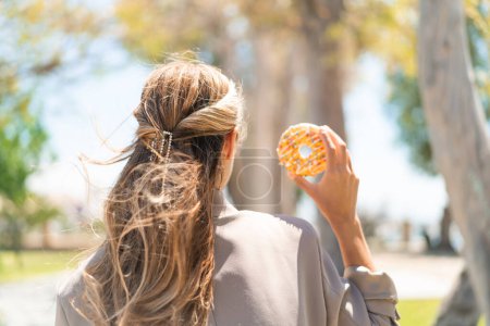 Photo for Pretty blonde Uruguayan woman holding a donut at outdoors in back position - Royalty Free Image