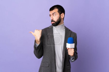 Téléchargez les photos : Adulte journaliste homme avec barbe tenant un microphone sur fond violet isolé malheureux et pointant vers le côté - en image libre de droit