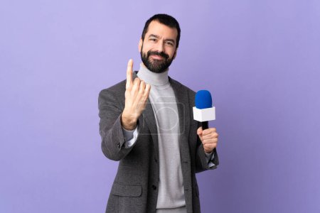 Téléchargez les photos : Reporter adulte homme avec barbe tenant un microphone sur fond violet isolé faisant geste à venir - en image libre de droit