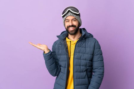 Téléchargez les photos : Skier homme avec des lunettes de snowboard sur fond violet isolé tenant copyspace imaginaire sur la paume pour insérer une annonce - en image libre de droit