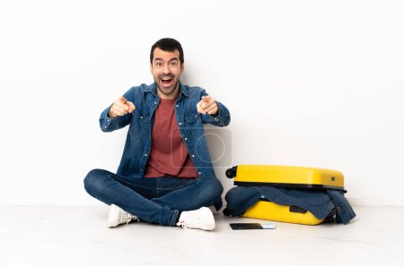 Foto de Caucásico hombre guapo con una maleta llena de ropa sentada en el suelo en el interior sorprendido y apuntando hacia delante - Imagen libre de derechos