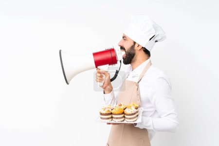 Téléchargez les photos : Jeune homme tenant gâteau muffin sur fond blanc isolé criant à travers un mégaphone - en image libre de droit