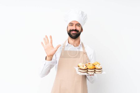 Téléchargez les photos : Jeune homme tenant gâteau muffin sur fond blanc isolé comptant cinq avec les doigts - en image libre de droit