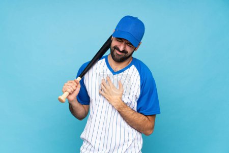 Junger Mann spielt Baseball vor isoliertem blauen Hintergrund und hat Herzschmerzen