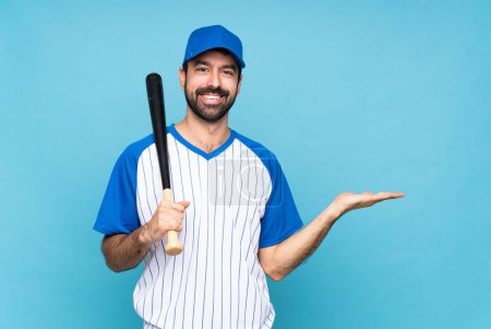 Téléchargez les photos : Jeune homme jouant au baseball sur fond bleu isolé tenant imaginaire copyspace sur la paume pour insérer une annonce - en image libre de droit