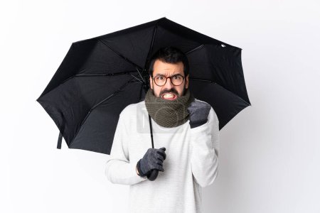 Foto de Hombre guapo caucásico con barba sosteniendo un paraguas sobre una pared blanca aislada frustrada por una mala situación - Imagen libre de derechos