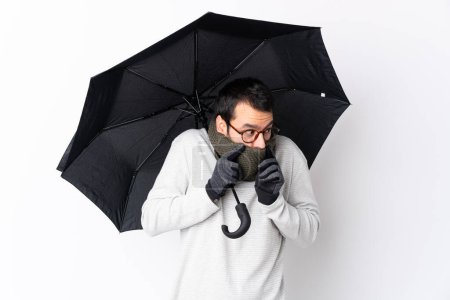 Foto de Hombre guapo caucásico con barba sosteniendo un paraguas sobre una pared blanca aislada - Imagen libre de derechos