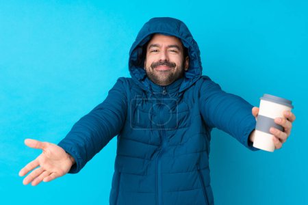 Téléchargez les photos : Homme vêtu d'une veste d'hiver et tenant un café à emporter sur fond bleu isolé présentant et invitant à venir avec la main - en image libre de droit