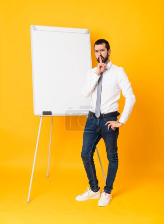 Téléchargez les photos : Plan complet de l'homme d'affaires faisant une présentation sur un tableau blanc sur fond jaune isolé montrant un signe de silence geste mettant le doigt dans la bouche - en image libre de droit