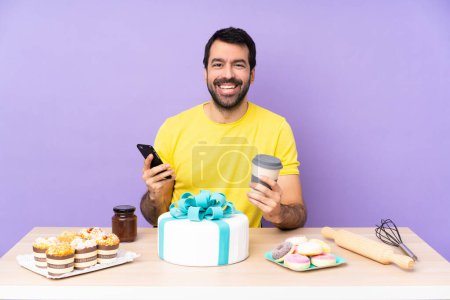 Foto de Hombre en una mesa con un gran pastel sosteniendo café para llevar y un móvil - Imagen libre de derechos