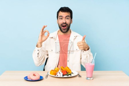 Foto de Hombre en una mesa desayunando gofres y un batido mostrando señal de ok y el pulgar hacia arriba gesto - Imagen libre de derechos