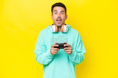 Foto de Joven hombre guapo jugando con un controlador de videojuegos sobre una pared aislada con expresión facial sorpresa - Imagen libre de derechos