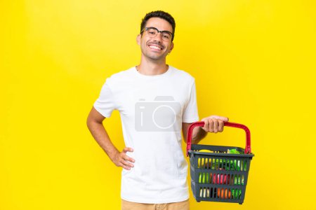 Photo for Young handsome man holding a shopping basket full of food over isolated yellow background posing with arms at hip and smiling - Royalty Free Image