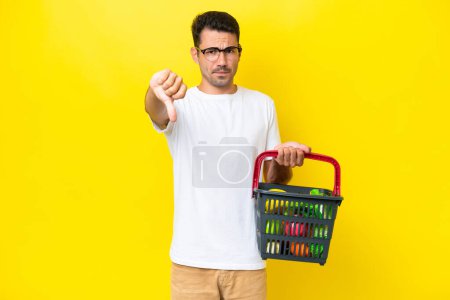 Téléchargez les photos : Jeune homme beau tenant un panier plein de nourriture sur fond jaune isolé montrant pouce vers le bas avec expression négative - en image libre de droit